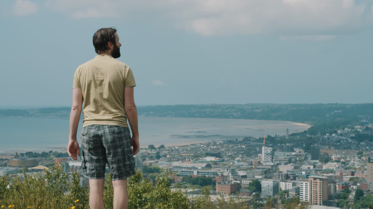 hombre en pantalones cortos y camiseta viendo el paisaje urbano durante una caminata en un día soleado en swansea, reino unido