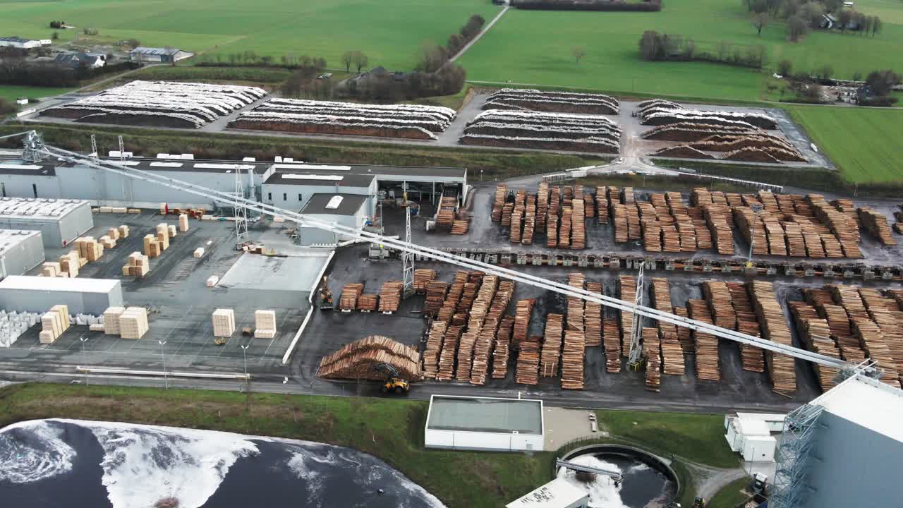 vista aérea de la fábrica de procesamiento de madera en alemania, troncos de madera de pino apilados en filas y maquinaria pesada en movimiento en el sitio
