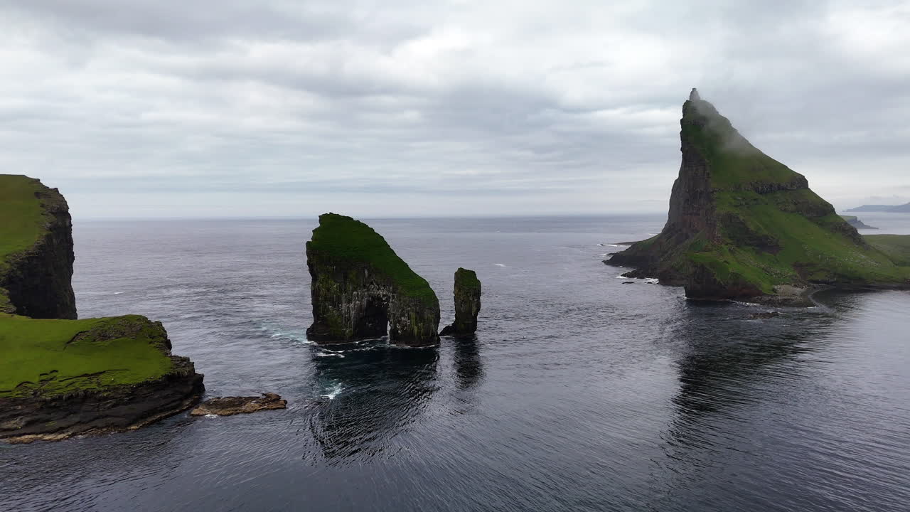 Cinematic aerial view of Drangarnir sea stacks rising dramatically from the Atlantic Ocean near Vágar, Faroe Islands, showcasing rugged cliffs, lush green slopes, and misty Nordic seascape