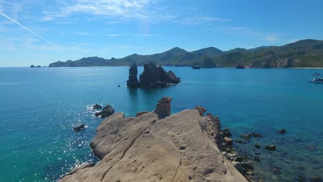 Flying-over-a-rocky-outcropped-peninsula-into-an-anchorage-with-passing-sailing-yacht-anchorage-in-the-turquoise-water-bay-in-Mexico