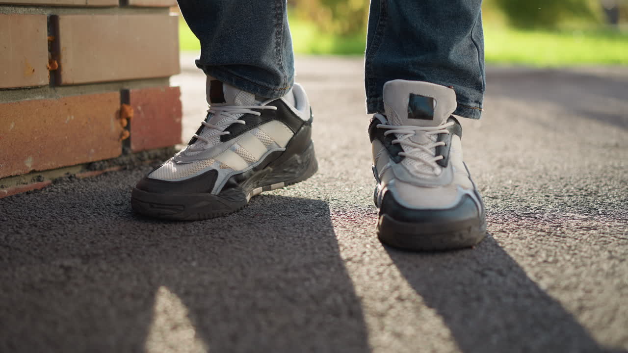 leg view of person wearing jeans and sneakers tapping right foot gently on asphalt surface beside brick wall under soft sunlight with shadows cast on pavement and blurred greenery in background