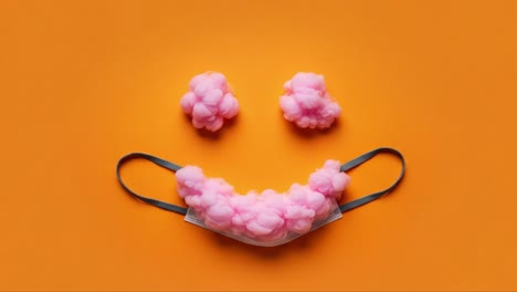 a creative arrangement of a face mask and pink clouds creating a cheerful expression against a vibrant orange background, symbolizing joy and positivity