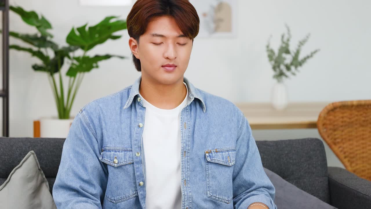 若いアジア人男性がソファの上であぐらをかき、目を閉じて瞑想している。
