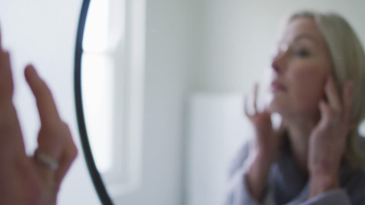 Donna anziana caucasica felice in bagno, che si guarda allo specchio e si tocca il viso.