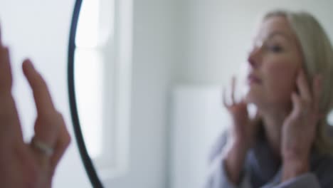 Happy-senior-caucasian-woman-in-bathroom,-looking-to-mirror,-touching-face