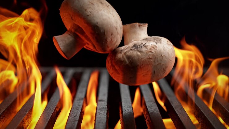 Mushrooms Fall Onto Charcoal Grill on the Black Background, Flames Burst From Beneath The Grill Grate in Slow Motion