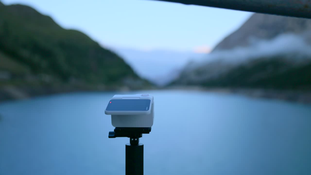 Una action camera Insta360 Go è montata su un treppiede con uno splendido lago alpino e montagne nebbiose sullo sfondo. Ideale per viaggi avventura, vlog e concetti di tecnologia moderna.