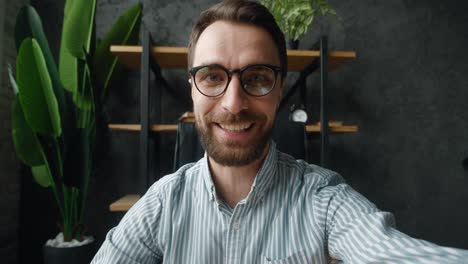 pov of handsome business man making selfie video call while sitting at modern office. portrait of a businessman blogger in eyeglasses holding smartphone, shooting video blog indoors at home office