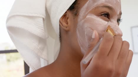 Happy-biracial-woman-with-towel-on-head-making-mask-on-face-in-bathroom,-slow-motion