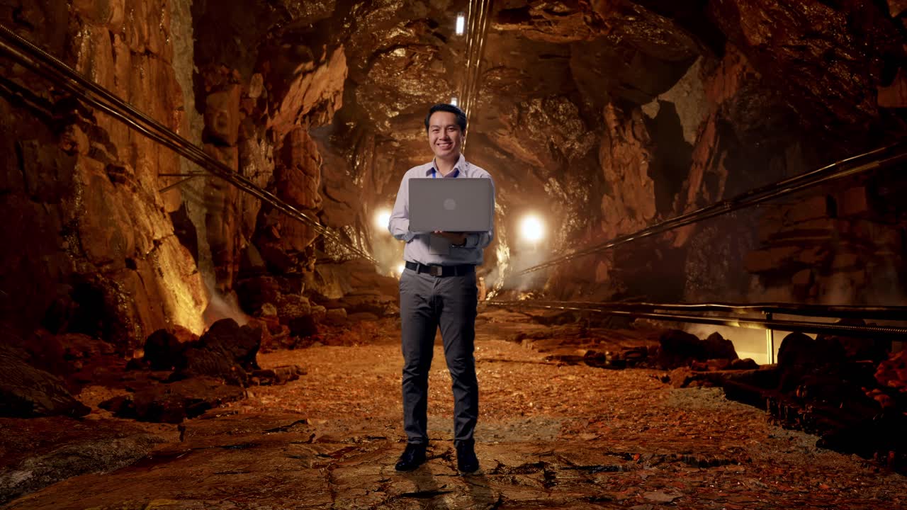 Full Body Of An Asian Male Professional Worker Standing With His Laptop In Underground Mine Tunnel, He Is Looking At The Camera With A Smile