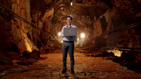 full body of an asian male professional worker standing with his laptop in underground mine tunnel, he is looking at the camera with a smile