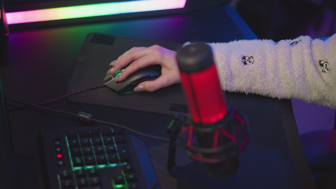 Lady with polished nail in fluffy sweater featuring teddy bear design using wired gaming mouse on black pad, surrounded by soft ambient lighting and streaming microphone, in cozy digital workstation