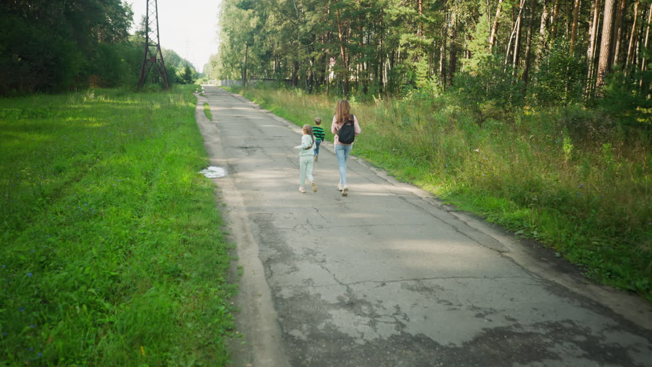 Mother and daughter joyfully follow young boy running ahead on cracked tarred countryside road, surrounded by lush greenery and tall trees under bright daylight