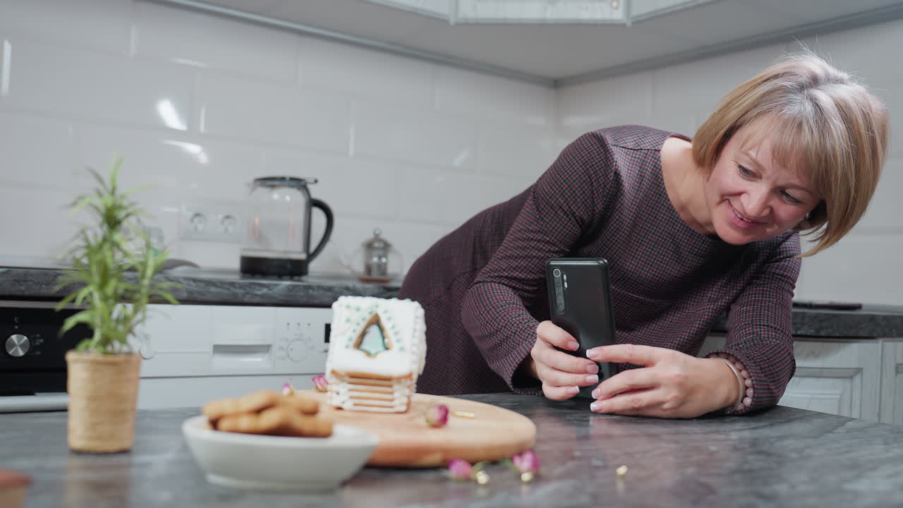 la panadera sonríe mientras toma una foto de su casa de pan de jengibre decorada usando un teléfono inteligente, la cocina acogedora presenta galletas, plantas en olla y equipos de cocina en el fondo