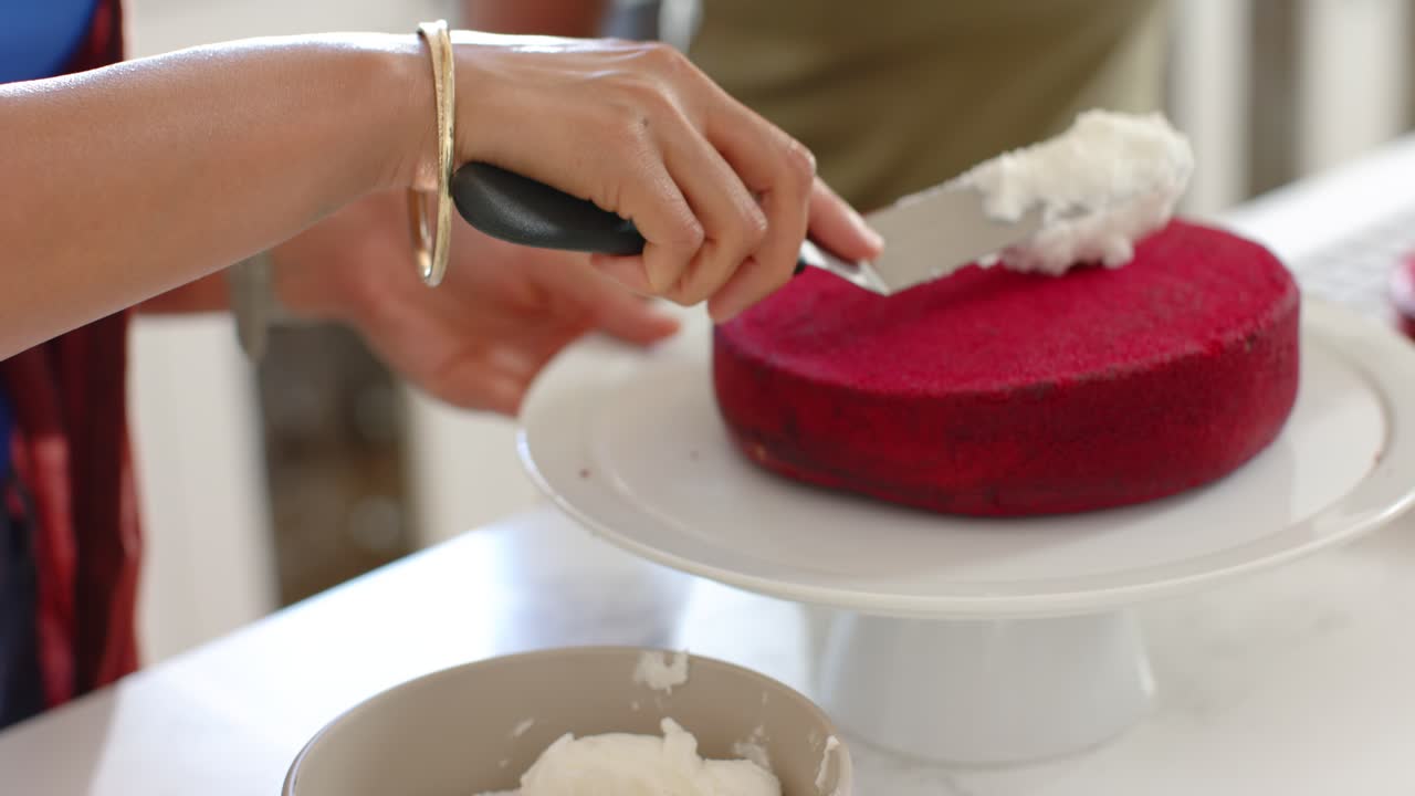Baker's hands are scooping, smoothing icing on red cake with helper steadying stand to frost cake