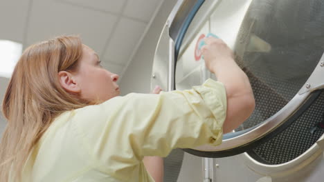 Lower view laundry worker in yellow shirt wiping wet glass surface of industrial washing machine with blue towel, showing soapy streaks across transparent door while focusing on cleanliness