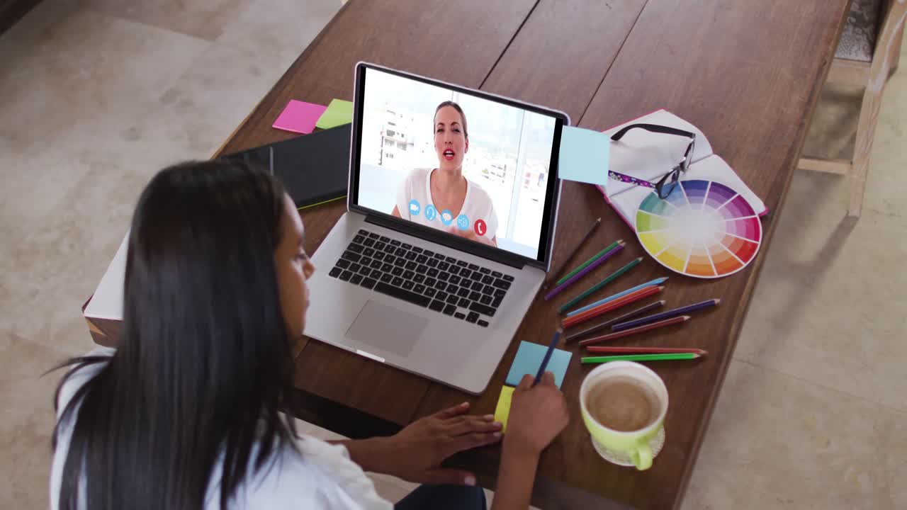 Mujer caucásica usando una laptop en videollamada con una colega, tomando notas