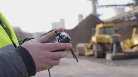 Man-controlling-a-drone-with-his-remote-controller,-filming-a-construction-site-in-a-yellow-vest