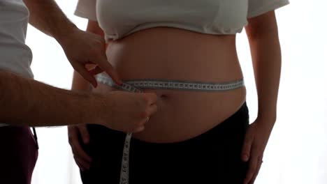 man measures pregnant woman's waist with tape while she raises arms and stands still