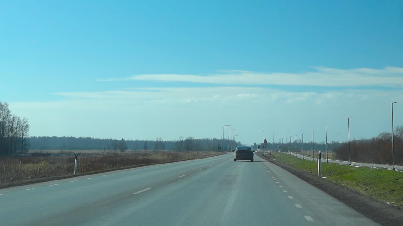 FPV or first person view from inside a car driving forward on a aspahalt road with white marking on a sunny summer day in Harku Estonia, blue sky, trees and farm fields visible on the sides and above.