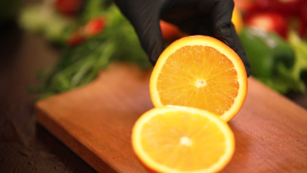 Arancia fresca per succo tagliata con un coltello grande su un tagliere da mani che indossano guanti neri con verdure sullo sfondo