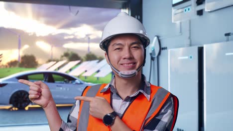 close up of asian male engineer with safety helmet smiling and pointing to side while standing with home energy storage system in a modern garage