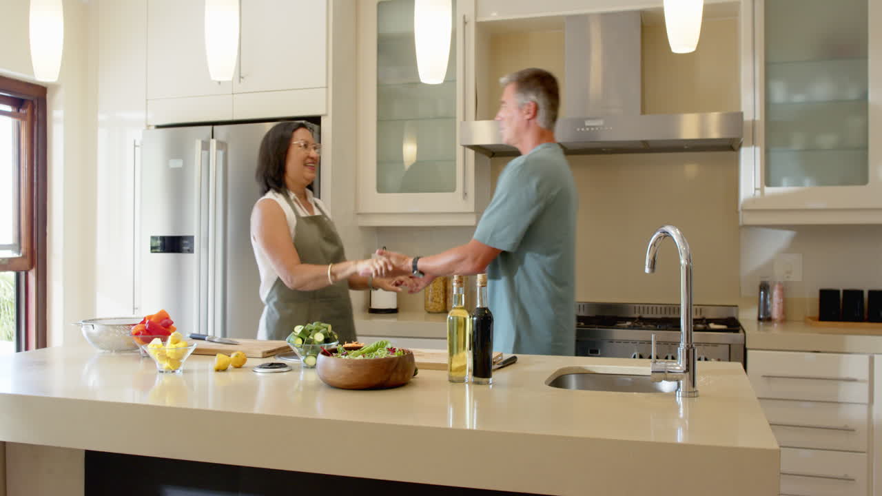 Casal idoso diverso se abraçando em cozinha moderna, aproveitando a manhã juntos em casa.