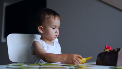 baby boy wearing white t-shirt focused on the game. serious kid sits at desk moving his toys.