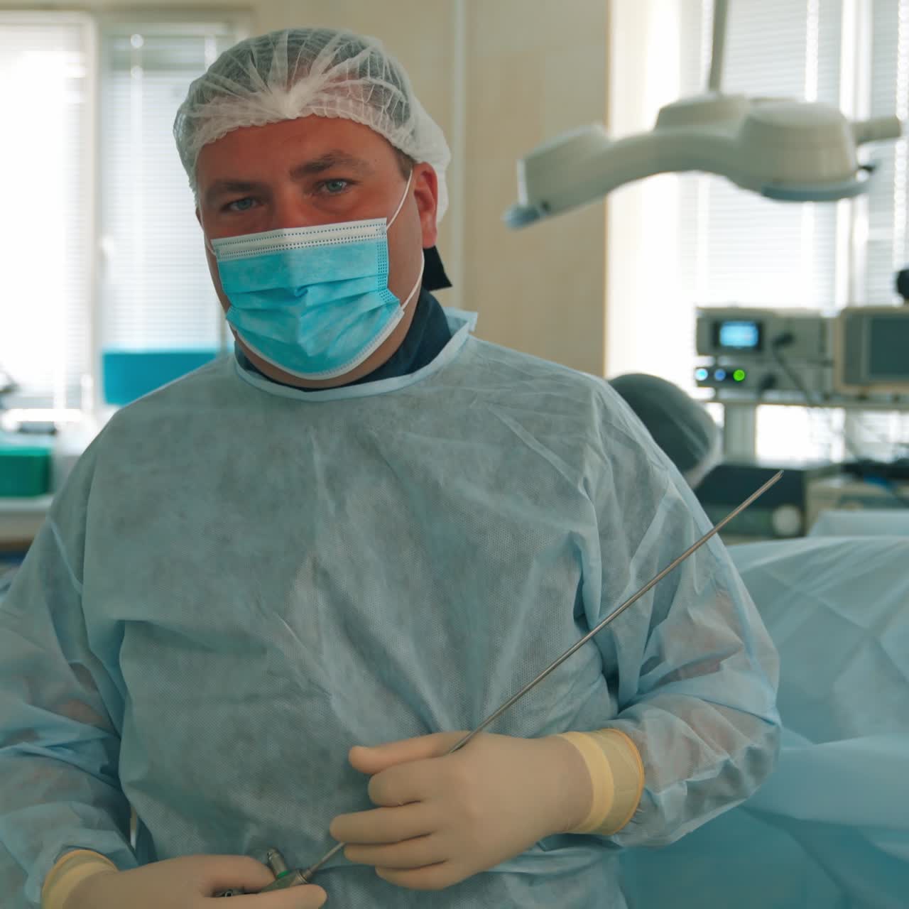 Medico di mezza età con mascherina e cappello, pronto per l'intervento, che parla alla telecamera. Chirurgo che tiene in mano un lungo ago con i guanti
