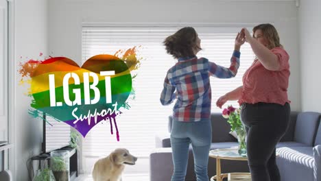 Animation-of-rainbow-heart-and-pride-over-lesbian-couple-dancing-at-home