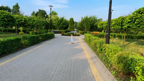 little baby boy wearing jeans romper is in the green park. active toddler runs by the road and stops.