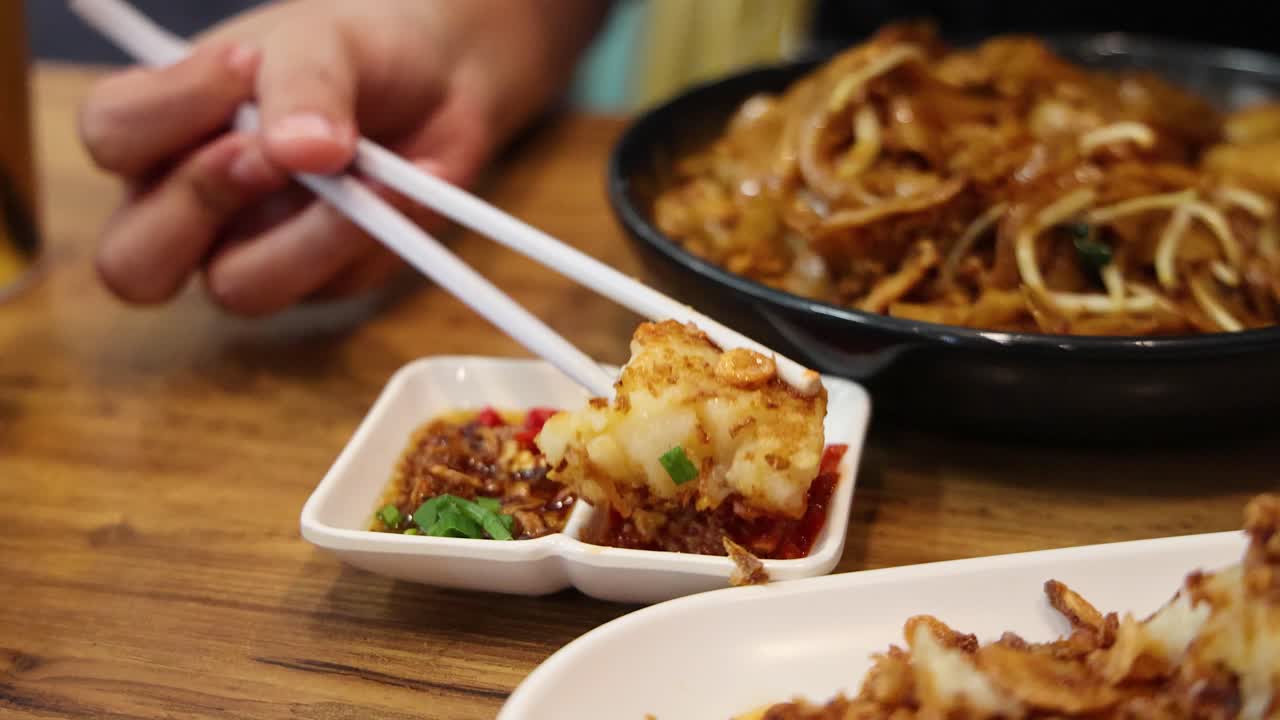 Hand uses chopsticks to dip a fried turnip cake into chili sauce, with noodles and dim sum on a wooden restaurant table under warm lighting