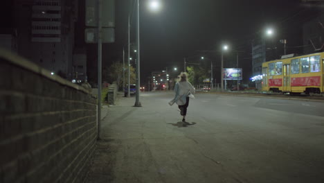 back view of woman in white coat running alone along empty city street at night under streetlights, casting long shadow on pavement as yellow tram moves past in calm urban setting
