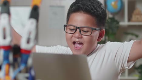 primer plano de un chico asiático lindo celebrando el éxito trabajando sobre una mano cyborg en una computadora portátil en casa