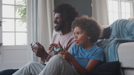 Padre-Negro-De-Mediana-Edad-Y-Su-Hijo-Preadolescente-Sentados-En-El-Suelo-De-Su-Sala-De-Estar-Jugando-Videojuegos-Juntos,-De-Cerca