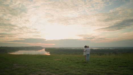 a couple stands on a grassy hill overlooking lake during sunset, shot from behind. the man, in a white shirt, hat, and jeans, holds the woman by the waist. she wears a black hat and a white dress