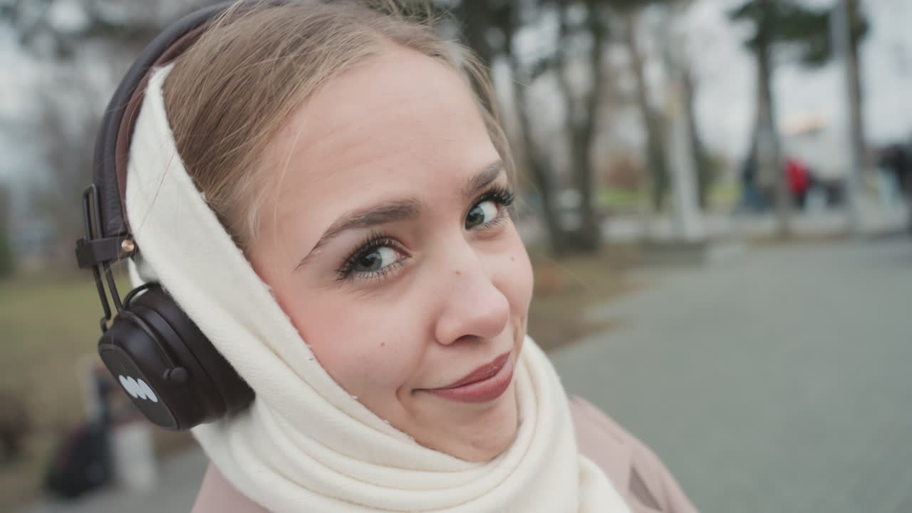 Primo piano: donna al parco che osserva attraverso un telescopio, con cuffie avvolgenti e sciarpa, un sorriso delicato e occhi concentrati. Ritratto intimo e spontaneo, con la texture della maglieria, alberi invernali e sfondo sfocato.