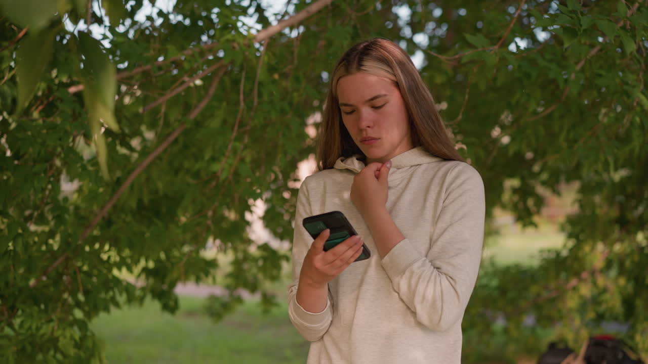 나무 에 서있는 여성, 드폰 화면에 깊이 집중하고, 손으로 후드 목걸이를 만지고, 잎을 통해 빛이 필터링되어 얼굴에 부드러운 그림자를 던지고, ბუნ적 배경이 흐릿합니다.