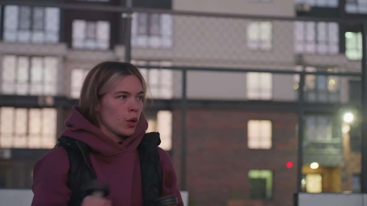 Gym goer boxing dumbbells punching air taking break mid workout on black asphalt sports court white barrier topped with chain link fence backdrop at dusk wearing maroon hoodie white sneakers