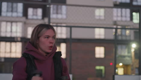 gym goer boxing dumbbells punching air taking break mid workout on black asphalt sports court white barrier topped with chain link fence backdrop at dusk wearing maroon hoodie white sneakers
