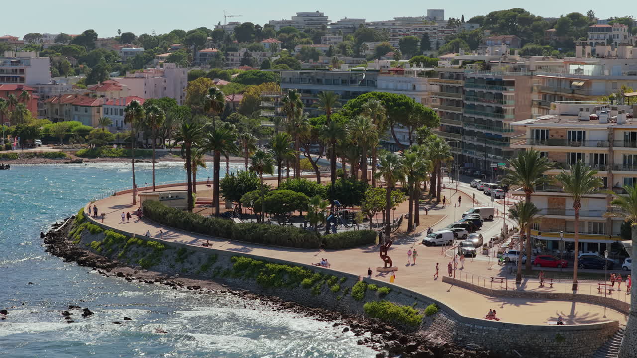 Aerial drone view of a coastal promenade lined with palm trees and public art sculptures, with people strolling near the Mediterranean Sea, Antibes, France