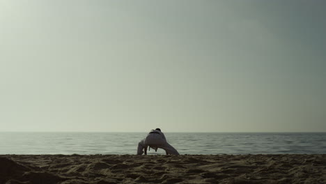 Karate-man-squat-stretching-legs-on-sandy-beach.-Athlete-training-flexibility.