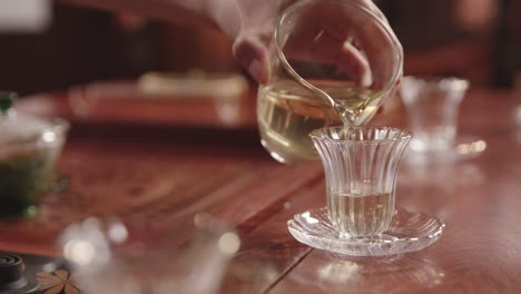 Chinese-woman-pours-freshly-prepared-green-tea-into-a-glass-cup-on-traditional-dark-wood-tea-table