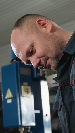 primer plano de un mecánico enfocado en un uniforme azul examinando algo fuera de la cámara en un taller automotriz con equipos de fondo borrosos