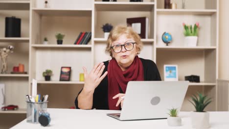 older woman in glasses sits at the table with a laptop, scolding while waving her hand and talking, looking at the camera