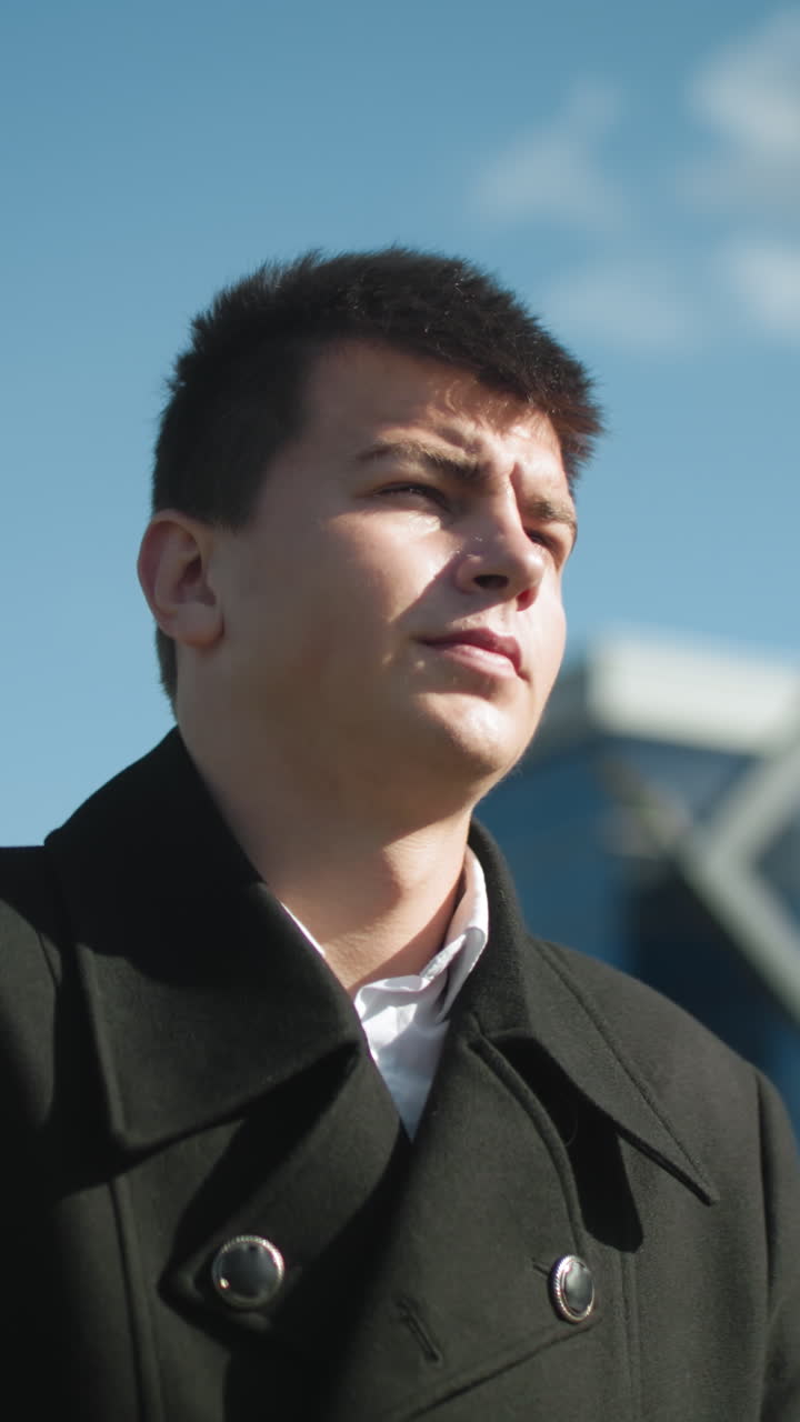 현대적인 유리 건물 근처의 도시 지역에서 밝은 빛 아래 야외를 고 있는 검은 코트를 입은 자신감 넘치는 사업가, 은 파란 하늘에 맞춰 단호하게 보입니다.