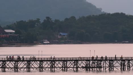 Mon-Bridge-and-a-longboat-followed-speeding-behind-the-structure-while-people-are-walking-on-the-bridge,-silhouetting-as-it-was-getting-dark,-in-slow-motion