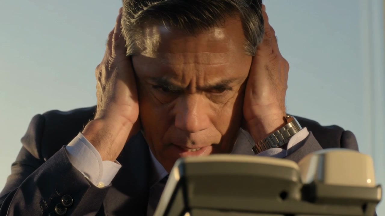 Un uomo in giacca e cravatta si china verso il telefono fisso che squilla, portandosi le mani alle orecchie per sentire, con un orologio da polso.