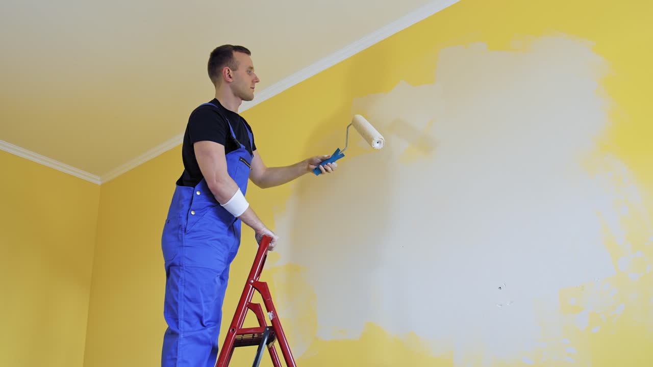 Worker repainting walls of the apartment from the ladder. Young builder male decorating walls into a new light color. Home repair.