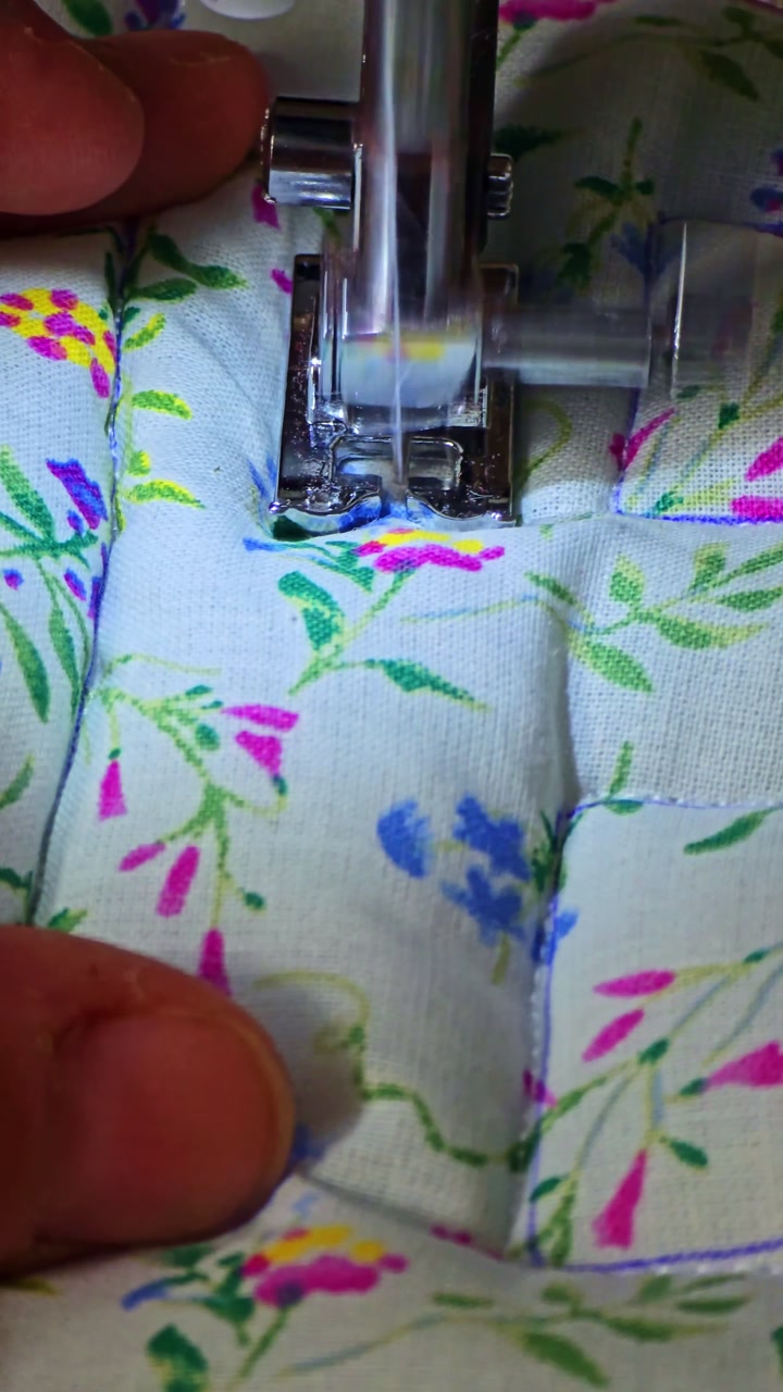 Close-up shot of a hand holding floral fabric while a sewing machine stitches, showing presser foot, thread, and fabric details.